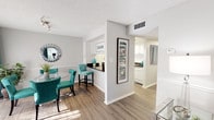 This thoughtfully designed single-floor residence offers a seamless blend of comfort and style, unified by a striking teal and white color scheme throughout. The open-concept living and dining area is anchored by warm hardwood-style flooring, chair-rail wainscoting, and abundant natural light streaming through large windows dressed in sheer curtains. The dining space features a glass-top round table surrounded by tufted teal chairs, complemented by a decorative sunburst mirror. A half-wall with a dark countertop bar connects the dining area to the galley-style kitchen, which is fitted with white cabinetry, dark countertops, a stainless steel sink, and white appliances. The living room offers a relaxed atmosphere with a neutral sofa accented by teal throw pillows and vibrant wall art. The primary bedroom features plush carpeting, warm wood furniture, and soft, neutral tones. A secondary bedroom provides versatile space with carpet flooring and a ceiling fan. The bathrooms are clean and bright, with wood-style flooring, white vanities, and tasteful décor. Generous closet space is provided throughout, with louvered bi-fold doors offering both style and practicality. The textured ceilings and consistent neutral wall tones tie the entire space together into a cohesive and inviting home.