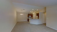 This single-floor residence offers a thoughtfully designed open-concept layout that connects the living, dining, and kitchen areas into one cohesive and functional space. The living area is finished with soft, neutral carpeting that adds warmth underfoot, while smooth walls painted in a light, airy tone create a bright and welcoming atmosphere. An elegant chandelier anchors the dining area, providing a refined focal point that complements the space's classic character.

The kitchen is a standout feature, showcasing rich medium-toned wood cabinetry that provides ample storage, paired with white appliances including a refrigerator, range, and microwave. A curved breakfast bar with a light countertop extends into the living area, offering a versatile space for casual dining or entertaining while maintaining visual continuity across the open floor plan. Recessed lighting and a ceiling fan with an integrated light fixture illuminate the kitchen and adjacent areas, ensuring the space is both practical and comfortable.

The overall ambiance is clean, neutral, and move-in ready, with a layout that maximizes flow and usability throughout the single level. The design strikes a balance between everyday functionality and a comfortable, livable aesthetic.