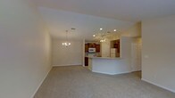 This single-floor residence offers a thoughtfully designed open-concept layout that connects the living, dining, and kitchen areas into one cohesive and functional space. The living area is finished with soft, neutral carpeting that adds warmth underfoot, while smooth walls painted in a light, airy tone create a bright and welcoming atmosphere. An elegant chandelier anchors the dining area, providing a refined focal point that complements the space's classic character.

The kitchen is a standout feature, showcasing rich medium-toned wood cabinetry that provides ample storage, paired with white appliances including a refrigerator, range, and microwave. A curved breakfast bar with a light countertop extends into the living area, offering a versatile space for casual dining or entertaining while maintaining visual continuity across the open floor plan. Recessed lighting and a ceiling fan with an integrated light fixture illuminate the kitchen and adjacent areas, ensuring the space is both practical and comfortable.

The overall ambiance is clean, neutral, and move-in ready, with a layout that maximizes flow and usability throughout the single level. The design strikes a balance between everyday functionality and a comfortable, livable aesthetic.