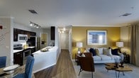 This single-story space showcases an open-concept design that seamlessly connects the kitchen and living area, creating a cohesive and functional flow throughout. The kitchen features dark cabinetry contrasted by a bright white countertop with an integrated sink, stainless steel appliances, and track lighting overhead that provides ample task illumination. A peninsula-style counter adds versatility, serving as both a prep space and a casual dining or gathering spot.

The living area is anchored by a warm golden-yellow accent wall, adding a welcoming pop of color to the otherwise neutral palette. Hardwood-style flooring runs throughout, lending a sense of continuity and warmth to the space. A hallway visible from the main living area leads toward additional rooms, hinting at a practical layout with good separation of spaces. Natural light filters in through a window in the living area, brightening the space alongside a stylish floor lamp. The combination of modern finishes, thoughtful lighting, and a cohesive color scheme gives this space a contemporary yet comfortable ambiance suited for everyday living.