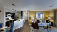 This single-story space showcases an open-concept design that seamlessly connects the kitchen and living area, creating a cohesive and functional flow throughout. The kitchen features dark cabinetry contrasted by a bright white countertop with an integrated sink, stainless steel appliances, and track lighting overhead that provides ample task illumination. A peninsula-style counter adds versatility, serving as both a prep space and a casual dining or gathering spot.

The living area is anchored by a warm golden-yellow accent wall, adding a welcoming pop of color to the otherwise neutral palette. Hardwood-style flooring runs throughout, lending a sense of continuity and warmth to the space. A hallway visible from the main living area leads toward additional rooms, hinting at a practical layout with good separation of spaces. Natural light filters in through a window in the living area, brightening the space alongside a stylish floor lamp. The combination of modern finishes, thoughtful lighting, and a cohesive color scheme gives this space a contemporary yet comfortable ambiance suited for everyday living.