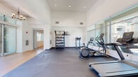 This well-appointed fitness center, located in Stockton, CA (95209), offers a clean, contemporary design with soaring ceilings and a crisp white interior that creates an open, energizing atmosphere. The main workout area is anchored by protective rubber flooring and equipped with a comprehensive selection of cardio machines, including treadmills, elliptical trainers, and a stationary bike. A full-length glass wall along one side of the room floods the space with natural light while offering views of a pool area beyond, making workouts feel connected to the outdoors. A dedicated free-weight and functional training zone features a multi-tiered storage rack stocked with kettlebells, dumbbells, resistance bands, and a stability ball, complemented by an overhead pull-up bar structure. A secondary corridor area with warm hardwood flooring transitions into a small lounge nook, where a stylish upholstered bench sits beneath a framed piece of colorful contemporary artwork, providing a welcoming rest area. Recessed lighting throughout ensures a well-lit environment at any time of day. The thoughtful layout and curated amenities make this fitness center a practical and attractive facility for daily use.