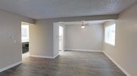 This single-story space presents an open and functional layout centered around a spacious living area. The flooring throughout is a wood-look laminate or vinyl plank in a warm gray-brown tone, providing a durable and cohesive foundation across the space. Neutral greige-toned walls complement the flooring, creating a calm, understated aesthetic that adapts well to a variety of furnishing styles.

The living area is defined by a textured popcorn ceiling and is equipped with a ceiling fan featuring an integrated light fixture, offering both air circulation and ambient lighting. A partial wall or soffit element divides portions of the open area, adding subtle architectural definition without fully closing off the space.

Natural light enters through a window on the far wall, brightening the room and contributing to an airy feel. White baseboards provide clean contrast against the neutral walls and flooring throughout.

An open doorway visible from the main living area leads to an adjacent room, likely a kitchen or utility space, where white appliances or cabinetry can be glimpsed, suggesting a practical and well-organized support area nearby. Overall, the space presents a move-in-ready, low-maintenance environment with a clean, modern-neutral character.