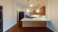 This single-floor residence in Minneapolis's Uptown neighborhood presents a thoughtfully designed open-concept layout that seamlessly connects the kitchen and living areas. The kitchen serves as the centerpiece of the space, showcasing a large waterfall-edge island with an undermount sink and striking globe pendant lighting suspended above. Warm wood cabinetry with glass-front upper panels complements the iridescent mosaic tile backsplash, while stainless steel appliances—including a French door refrigerator, wall oven, and range hood—complete the upscale culinary setup. Rich, dark hardwood flooring flows throughout the main living areas, adding warmth and continuity. The living area opens directly onto a private outdoor balcony through full-height sliding glass doors, allowing natural light to pour in and providing outdoor views of the surrounding urban landscape. A bedroom features plush carpeting, a ceiling fan, and a bright window. The bathroom offers a clean, contemporary aesthetic with a large walk-in shower fitted with sliding glass doors, mosaic tile flooring, and a sleek vanity with an integrated sink. Neutral wall tones and modern finishes throughout contribute to a cohesive, contemporary ambiance.