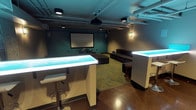 Located at 4040 26th Ave SW in Seattle, WA, this single-floor entertainment lounge is thoughtfully designed for social gatherings and cinematic experiences. The space centers around a dedicated screening area anchored by a large projection screen mounted against a bold teal accent wall, complemented by a ceiling-mounted projector and flanking wall-mounted speakers that create an immersive viewing environment. Generous modular dark gray sectional seating and ottomans are arranged on a patterned area rug, offering ample room for a crowd to gather comfortably. Exposed ductwork and structural elements run across the ceiling, lending the space an industrial aesthetic that is softened by ambient wall sconces casting a warm glow throughout. Two illuminated bar-height counters with glowing blue-green light panels serve as striking focal points, paired with sleek chrome bar stools for casual seating. A wet bar area along one wall features a stainless steel sink and cabinetry, providing practical support for entertaining. The polished aggregate concrete flooring ties the industrial and modern design elements together seamlessly. The overall ambiance is cool, moody, and well-suited for private screenings, social events, or community use.