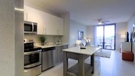 This single-floor residence offers a thoughtfully designed open-concept layout that seamlessly connects the kitchen, dining, and living areas. The contemporary kitchen is equipped with stainless steel appliances, including a range, microwave, and dishwasher, complemented by crisp white cabinetry with modern hardware and light-toned countertops. A freestanding kitchen island provides additional prep space and casual seating, making it a functional centerpiece for everyday living and entertaining. The space is bathed in natural light, streaming in through floor-to-ceiling glass doors that lead to a private balcony, offering open outdoor views and a seamless indoor-outdoor connection. A ceiling fan in the living area promotes air circulation and adds a subtle architectural detail overhead. The light-toned flooring throughout ties the spaces together, enhancing the bright and airy ambiance. The overall design aesthetic is clean, minimalist, and modern, with a neutral color palette that creates a calm and welcoming atmosphere throughout the residence.