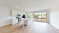 Located at 1115 Seigle Ave in Charlotte's vibrant 28205 zip code, this single-floor residence offers a thoughtfully designed open-concept layout finished with contemporary details throughout. Light wood-tone flooring runs continuously across the entire unit, creating a cohesive and airy feel from room to room.

The kitchen serves as a central highlight, featuring crisp white shaker cabinetry, a classic white subway tile backsplash, and stainless steel appliances including an electric range, microwave, and French door refrigerator. A large central island with a white countertop and an undermount sink provides generous prep space and is accented by modern pendant lighting. Expansive wood-framed windows flood both the kitchen and adjacent living area with natural light and offer views of the surrounding greenery.

The open living area flows seamlessly from the kitchen, providing a flexible and well-lit space. Multiple bedrooms are positioned along a connecting hallway, each equipped with wood-framed windows that bring in ample daylight. The unit includes two full bathrooms — one featuring a walk-in glass shower stall and the other a full bathtub — both finished with white shaker vanities and dark countertops. Clean white walls and consistent flooring throughout lend the entire space a bright, modern, and move-in-ready character.