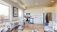 This thoughtfully designed single-floor residence offers a bright and contemporary living experience. The open-concept layout seamlessly connects the kitchen, dining, and living areas, creating an inviting and functional space. The kitchen is outfitted with crisp white cabinetry extending to the ceiling, a classic white subway tile backsplash, light stone countertops, and a full suite of stainless steel appliances, including a range, microwave, dishwasher, and refrigerator. The dining area sits adjacent to the kitchen, bathed in natural light from large windows that frame expansive views of the surrounding landscape. The living area continues the bright, airy aesthetic with warm light wood flooring that flows throughout the entire residence. A dedicated office or den features floor-to-ceiling windows offering sweeping outdoor views, making it an ideal workspace. The bedroom is a serene retreat, highlighted by a large window with scenic vistas. The full bathroom features white shaker-style cabinetry, a clean white vanity, and a tub-shower combination. A clean, well-lit hallway connects the private rooms to the main living space. Recessed lighting and neutral warm tones throughout contribute to the residence's modern and cohesive ambiance.