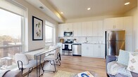 This thoughtfully designed single-floor residence offers a bright and contemporary living experience. The open-concept layout seamlessly connects the kitchen, dining, and living areas, creating an inviting and functional space. The kitchen is outfitted with crisp white cabinetry extending to the ceiling, a classic white subway tile backsplash, light stone countertops, and a full suite of stainless steel appliances, including a range, microwave, dishwasher, and refrigerator. The dining area sits adjacent to the kitchen, bathed in natural light from large windows that frame expansive views of the surrounding landscape. The living area continues the bright, airy aesthetic with warm light wood flooring that flows throughout the entire residence. A dedicated office or den features floor-to-ceiling windows offering sweeping outdoor views, making it an ideal workspace. The bedroom is a serene retreat, highlighted by a large window with scenic vistas. The full bathroom features white shaker-style cabinetry, a clean white vanity, and a tub-shower combination. A clean, well-lit hallway connects the private rooms to the main living space. Recessed lighting and neutral warm tones throughout contribute to the residence's modern and cohesive ambiance.