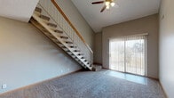 This two-floor space is defined by a dramatic vaulted ceiling in the main living area, where an open staircase with a wood-rail banister and carpeted treads serves as a central architectural feature. A ceiling fan is mounted at the peak of the vaulted ceiling, and a sliding glass door fitted with vertical blinds provides outdoor access and fills the living space with natural light.

The kitchen is laid out in a galley-style configuration, featuring oak cabinetry with brushed chrome hardware, dark granite-style countertops, tile flooring, and black appliances including a full-size refrigerator. Storage is well-distributed with both upper and lower cabinets.

Two bathrooms are present, each finished with oak cabinetry, dark countertops, tile flooring, and a tub/shower combination. One bathroom includes a tall storage cabinet alongside the vanity.

The upper level opens to a spacious landing overlooking the staircase and includes a room with a skylight that provides ample natural illumination. Additional carpeted rooms feature neutral-toned walls, wood baseboards, and windows with horizontal blinds, offering versatile spaces throughout both floors.