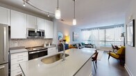 This single-floor residence showcases a contemporary open-concept design that seamlessly integrates the kitchen, dining, and living areas into one cohesive, light-filled space. The kitchen is a standout feature, equipped with stainless steel appliances including a gas range, microwave, and refrigerator. White shaker cabinetry pairs elegantly with a neutral tile backsplash, while a generous island with a quartz countertop and undermount sink serves as the centerpiece, topped by sleek pendant lighting. Track lighting along a curved rail adds additional illumination throughout the cooking space.

Wood-look flooring flows throughout the main living area, which is anchored by floor-to-ceiling windows that flood the space with natural light and offer outdoor views. The living room provides a comfortable, well-proportioned layout suited for both relaxing and entertaining.

The bedroom features plush carpeting, a glass door providing natural light and outdoor access, and dual sliding-door closets offering generous storage. A private bathroom sits conveniently adjacent. A dedicated in-unit laundry area houses a stacked washer and dryer, adding practical convenience. Throughout, the design maintains a clean, modern aesthetic with a neutral palette and thoughtful finishes.
