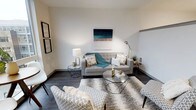 This single-floor space showcases a thoughtfully designed living area that combines modern aesthetics with functional comfort. The room features dark hardwood flooring that contrasts elegantly with the crisp white walls, creating a clean and polished atmosphere throughout.

The living area is anchored by a light grey tufted sofa adorned with decorative pillows and a teal throw blanket, complemented by a pair of patterned accent chairs that add visual interest to the space. A geometric glass-top coffee table serves as a central focal point, reflecting light and contributing to the room's contemporary character.

Wall décor includes a coastal-inspired canvas painting above the sofa and a pair of framed prints along the adjacent wall, adding personality without overwhelming the neutral palette. Layered area rugs provide texture and warmth underfoot, while a sleek brass floor lamp and a table lamp offer versatile ambient lighting options.

Side tables with geometric designs echo the modern design language found throughout the space. The overall ambiance is bright, airy, and inviting, making effective use of clean lines and a cohesive color scheme of whites, greys, and teal accents.