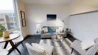 This single-floor space showcases a thoughtfully designed living area that combines modern aesthetics with functional comfort. The room features dark hardwood flooring that contrasts elegantly with the crisp white walls, creating a clean and polished atmosphere throughout.

The living area is anchored by a light grey tufted sofa adorned with decorative pillows and a teal throw blanket, complemented by a pair of patterned accent chairs that add visual interest to the space. A geometric glass-top coffee table serves as a central focal point, reflecting light and contributing to the room's contemporary character.

Wall décor includes a coastal-inspired canvas painting above the sofa and a pair of framed prints along the adjacent wall, adding personality without overwhelming the neutral palette. Layered area rugs provide texture and warmth underfoot, while a sleek brass floor lamp and a table lamp offer versatile ambient lighting options.

Side tables with geometric designs echo the modern design language found throughout the space. The overall ambiance is bright, airy, and inviting, making effective use of clean lines and a cohesive color scheme of whites, greys, and teal accents.