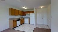 This single-story space offers a practical and well-organized layout designed for comfortable everyday living. The kitchen serves as a central feature, equipped with warm honey-toned wood cabinetry that provides generous storage across both upper and lower cabinets. White appliances, including a refrigerator, electric range, and dishwasher, complement the clean, neutral color palette throughout. A built-in range hood sits above the stove, and the countertops offer ample workspace for meal preparation. The kitchen sink is positioned along the counter with a functional layout that maximizes efficiency. The flooring in the kitchen and entry area consists of light-colored square tiles, creating a clean and easy-to-maintain surface. A transition to carpet is visible toward the adjacent living area, adding a sense of warmth and distinction between spaces. The main entry door is positioned just off the kitchen, featuring a solid white panel door with visible hardware, providing a straightforward and accessible entryway. The ceilings feature a subtle texture finish, and overhead fluorescent and globe lighting ensure the space is well-lit. Overall, the space presents a no-frills, functional design suited for practical day-to-day use.