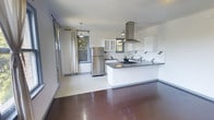 This single-floor space showcases an open-plan layout that seamlessly connects the living area and kitchen, creating a cohesive and functional environment. The kitchen is a standout feature, equipped with white cabinetry — including glass-front upper cabinets — and dark countertops that provide a striking contrast. A stainless steel range hood serves as a focal point above the gas cooktop, complemented by a stainless steel refrigerator and a dedicated prep sink, making the space well-suited for cooking enthusiasts. Decorative pendant lighting adds warmth and character overhead.

The flooring transitions between polished dark concrete in the living area and large-format light tiles in the kitchen zone, clearly delineating the two spaces while maintaining a unified aesthetic. White walls throughout keep the interior bright and clean. Multiple windows dressed with neutral linen curtains invite generous natural light and offer views of surrounding greenery, enhancing the sense of openness. The overall design balances modern minimalism with practical functionality, resulting in a bright, well-appointed space suitable for everyday living.
