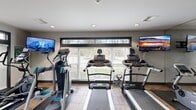 This fitness center is organized across two connected rooms, offering a comprehensive range of exercise options within a functional and well-maintained layout. The main cardio area is bathed in natural light from a broad horizontal window that stretches across the wall, providing pleasant views of the surrounding greenery. This room houses multiple treadmills, elliptical trainers, and an upright stationary bike, all positioned to take advantage of the outdoor view. Wall-mounted flat-screen TVs are strategically placed throughout for entertainment during workouts. Recessed lighting and neutral gray walls contribute to a clean, modern aesthetic, while durable tile flooring runs throughout the space.

The secondary room is dedicated to strength training and features a multi-functional cable machine, a squat rack with free weights, weight benches, a dumbbell rack, and accessory items such as a stability ball and balance board. Floor-to-ceiling mirrors line one wall, enhancing the sense of space and allowing users to monitor their form. Charcoal-framed glass panel doors connect the two areas, adding a polished architectural detail. Both rooms are well-lit with a combination of recessed and overhead fluorescent fixtures, ensuring a bright and energizing environment suitable for a full range of workout routines.