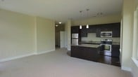 This single-floor space showcases an open-concept layout that seamlessly connects the kitchen and living areas. The kitchen serves as a focal point, featuring rich dark-espresso cabinetry that extends to the upper walls, providing generous storage throughout. Granite countertops add a touch of elegance, while the full suite of stainless steel appliances — including a refrigerator, stove, and built-in microwave — lends a polished, contemporary feel. A curved kitchen island with a built-in sink offers additional prep space and doubles as a casual dining or gathering spot. Three pendant lights hang above the island, providing targeted task lighting and a modern aesthetic detail. The transition from the kitchen's hard flooring to the plush neutral carpeting in the living area subtly defines each zone while maintaining an open, airy atmosphere. Soft, neutral-toned walls throughout the space reflect light evenly, contributing to a clean and welcoming ambiance. A hallway visible in the background leads to additional areas of the unit, hinting at a thoughtfully organized floor plan designed for comfortable everyday living.