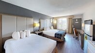 Located at 1400 West Tunnel Blvd in Houma, Louisiana, this hotel room offers a practical and comfortable layout designed to accommodate guests with a range of needs. The main sleeping area features two queen-sized beds dressed in crisp white linens, each backed by tall upholstered headboards set against a contrasting dark accent wall. Warm wood-look flooring runs throughout, complemented by neutral tones and tasteful wall art that create a calm, welcoming atmosphere. Natural light filters in through a large window dressed with curtain panels, further enhancing the room's open feel.

A dedicated workspace includes a desk and ergonomic chair positioned alongside a dresser with a flat-screen television, making the room well-suited for business travelers. A compact kitchenette provides added convenience, equipped with a mini refrigerator, microwave, stovetop, and sink with cabinet storage — practical amenities for extended stays.

The room includes two bathrooms, offering added privacy and convenience for multiple guests. One bathroom features a full tub-and-shower combination with warm-toned tile surround, a toilet, vanity, and wall-mounted hair dryer. The second bathroom area includes a granite-style countertop sink, an open wardrobe with hanging rack, and additional toiletry storage. Together, these spaces provide a functional, comfortable base for exploring the Houma area.
