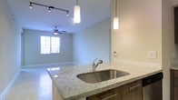 This well-appointed single-floor residence showcases a thoughtfully designed open-concept layout that seamlessly blends the kitchen and living areas into one cohesive, airy space. The kitchen is a standout feature, anchored by a granite-topped island with subtle speckling that catches the light beautifully. The island is equipped with a stainless steel undermount sink and pull-out faucet, complemented by rich, dark cabinetry that adds warmth and contrast. Pendant lighting hangs gracefully above the island, providing focused illumination and a touch of modern elegance. A track lighting system extends across the ceiling, offering flexible, adjustable light throughout the space. The living area flows naturally from the kitchen, featuring light-toned tile flooring that enhances the sense of openness and cleanliness. A ceiling fan with integrated lighting anchors the living space, contributing both comfort and style. Natural light filters in through a window fitted with blinds, brightening the room and connecting the interior to the outdoors. Neutral wall tones throughout create a calm, versatile backdrop suited to a variety of décor styles. A wall-mounted thermostat adds a practical, modern touch to the overall design of this inviting and functional home.