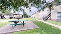 This single-story residential community is thoughtfully designed around a spacious, open courtyard that serves as the heart of the property. The grounds are well-maintained, featuring neatly trimmed green lawns that stretch between the building structures, creating an inviting and open atmosphere. Mature trees provide natural shade throughout the outdoor areas, enhancing comfort during warmer months.

The outdoor common area includes a picnic table with a dedicated concrete pad, ideal for casual dining and socializing. A charcoal grill positioned nearby adds to the outdoor entertainment options available to residents. A covered gazebo is visible in the background, offering a sheltered gathering space that complements the overall outdoor amenity offerings.

The buildings themselves present a clean, neutral exterior finish with green-accented rooflines that give the property a cohesive and tidy appearance. Exterior metal staircases provide access to upper-level units, while ground-floor entries open directly to the courtyard. The layout of the buildings creates a sense of community while maintaining open sightlines across the property. The generous green space between structures ensures residents enjoy both privacy and a welcoming shared environment, making this a functional and pleasant place to call home.