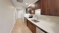 This single-floor residence offers a practical and well-designed layout across two bedrooms, one bathroom, a living room, kitchen, and a closet. The living room is a standout feature, bathed in natural light through a trio of large, white-framed sash windows that frame views of the surrounding urban streetscape, complemented by light hardwood flooring and recessed lighting overhead. The galley-style kitchen is efficiently designed with rich dark wood cabinetry, light quartz countertops, a stainless steel sink, and a full suite of stainless steel appliances including a range, microwave, and refrigerator. A long corridor connects the kitchen to the rest of the unit, reinforcing the linear flow of the layout. The primary bedroom features light hardwood flooring, a ceiling fan, and a window allowing natural light, while the second bedroom is carpeted with neutral tones and includes a ceiling fan, a window, and a double-door closet. The bathroom is finished with classic white subway tile surrounding the tub-shower combination, marble-look tile flooring, a vanity with dark wood cabinetry, and a frameless mirror. Throughout, neutral warm-beige walls and clean white trim create a cohesive, modern aesthetic.