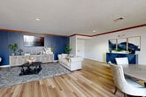 Located at 17131 Frances Plaza in Omaha, Nebraska, this single-floor space offers a generous and versatile layout designed for comfortable gathering and socializing. The interior is unified by light-toned hardwood flooring that extends throughout, complemented by crisp white walls adorned with tasteful crown molding that adds a refined architectural touch.

The space comprises two distinct living areas, each with its own character. The first is a lounge-style room anchored by a bold navy blue accent wall, a wall-mounted flat-screen television, and a pair of tufted cream sofas arranged around a dark wood coffee table atop a patterned area rug. A coordinating navy geometric sideboard and neutral media console complete the sophisticated, curated aesthetic. The second living area takes on a more open, social atmosphere, featuring multiple round dining tables surrounded by upholstered chairs, ideal for group dining or meeting settings. Natural light filters in through a large window, brightening the room and highlighting the clean, airy ambiance. Recessed lighting and crown molding with warm wood accents are consistent throughout both areas.

An elevator provides convenient access, and a closet with elegant gold hardware offers practical storage. The overall design balances modern style with functional versatility, making this a welcoming and well-appointed communal space.
