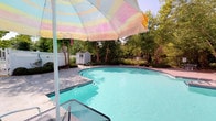 This outdoor pool area offers a relaxing retreat centered around a generously sized freeform pool with a classic kidney-inspired shape and clear, turquoise water. The pool features built-in entry steps and a metal handrail ladder, providing convenient access at multiple points. The surrounding deck is composed of light-toned pavers, offering ample space for lounge furniture and shade structures. A colorful striped patio umbrella provides a cheerful focal point and practical sun protection for poolside seating. The area is enclosed by a white vinyl privacy fence, creating a secluded and secure environment. A compact storage shed situated near the pool provides practical space for pool equipment and accessories. Mature deciduous trees line the perimeter, offering natural shade and a lush green backdrop that enhances the private, park-like atmosphere. Landscaped planting beds along the fence line add additional greenery and visual interest. The overall ambiance is bright, open, and inviting, making it a well-appointed outdoor space suited for warm-weather relaxation and recreation.