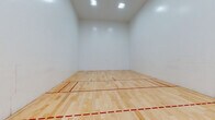 This dedicated racquetball facility is purpose-built for competitive and recreational play, offering a well-maintained, regulation-sized court designed to meet professional standards. The court features a polished hardwood floor with clearly marked red boundary and service lines, providing excellent traction and rebound consistency essential for the sport.

The three playing walls are finished in smooth, bright white — a hallmark of proper racquetball court construction — allowing players to track the ball with ease against a clean, unobstructed surface. The court benefits from overhead recessed lighting that evenly illuminates the entire playing area, ensuring optimal visibility during play.

A standout feature of this facility is the full-height glass back wall, composed of frameless glass panels with minimal hardware, offering complete transparency into the court. Adjacent to the glass wall is a carpeted viewing and waiting area, elevated slightly above the court level, with tiered seating for spectators or players awaiting their turn. This viewing area is generously sized and benefits from natural light via windows that overlook the exterior.

The overall design is clean, functional, and professional, reflecting a serious commitment to the sport. The combination of high ceilings, bright surfaces, quality flooring, and a dedicated spectator area makes this a well-rounded racquetball facility suited for both casual players and competitive athletes.