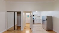 This single-floor space offers a clean, well-maintained layout with a cohesive design throughout. Light hardwood flooring runs continuously across all areas, creating a warm and unified aesthetic that enhances the sense of openness.

The main living area is generously proportioned, featuring crown molding, a ceiling fan with integrated lighting, and a wide window that frames lush greenery outside, filling the room with natural light. A door provides convenient exterior access directly from this space.

Adjacent to the main room, the kitchen is efficiently designed with white shaker-style cabinetry, light speckled stone countertops, and a matching tile backsplash. Stainless steel appliances — including a gas range, over-the-range microwave, and a full-size refrigerator — complete the functional setup.

The sleeping area includes a full-width mirrored sliding closet, offering ample storage while visually expanding the room. Crown molding continues here, maintaining the consistent architectural detailing found throughout the unit.

The bathroom is finished in a bright, all-white palette with classic square wall tiles surrounding a bathtub and shower combination, complemented by bronze fixtures. A white vanity with a lighted mirror and a window adds both practicality and a touch of natural light to the space.