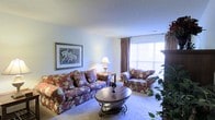 This single-floor residence offers a comfortable and classically styled living environment. The living room serves as the heart of the space, featuring plush wall-to-wall carpeting that adds warmth underfoot. A large window dressed with elegant drape curtains floods the room with natural light, creating a bright and welcoming ambiance throughout the day. The room is thoughtfully arranged with a floral-upholstered sofa and matching armchair, centered around a decorative oval coffee table with ornate metal legs — lending the space a traditional, homely character. A framed botanical artwork adorns the wall, adding a touch of personality and charm. Soft, neutral-toned walls complement the warm tones of the furnishings, while a classic table lamp provides additional ambient lighting. Lush indoor greenery near the window further enhances the inviting atmosphere of the room. The overall aesthetic of the space is cozy and well-maintained, with a timeless residential style that emphasizes comfort and livability. This single-story layout ensures easy accessibility and a seamless flow throughout the home.