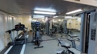 This fitness center offers a practical and fully equipped environment for a wide range of workout routines. The space is outfitted with a variety of cardio machines, including treadmills and elliptical trainers, positioned for easy access and use. A selection of strength training machines and cable systems occupies the central area, providing options for full-body resistance training. The flooring features durable, textured carpet or rubber matting suited for high-traffic athletic use. Floor-to-ceiling mirrors line the walls, visually expanding the space and allowing users to monitor their form during exercise. Overhead fluorescent lighting ensures the entire area is well-lit, contributing to a functional and focused atmosphere. The layout is thoughtfully organized to allow users to move between equipment with ease, maximizing the usable floor space. The overall ambiance is clean and utilitarian, designed with purpose and efficiency in mind, making it a functional facility for both cardiovascular conditioning and muscle strengthening activities.