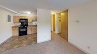 This 1-bedroom, 1-bathroom unit is thoughtfully designed to maximize functionality across a single floor. The kitchen is equipped with light maple cabinetry offering generous storage, dark granite-style countertops, a double-basin sink, and black appliances including an electric range, built-in microwave, and oven. Stone-look tile flooring grounds the kitchen space and provides durability in this high-traffic area. The kitchen opens toward the living area, which is finished with neutral carpet and features a textured ceiling and crown molding details. A wall-mounted air conditioning unit and baseboard heating ensure year-round comfort. Windows dressed with vertical blinds bring in natural light and offer outdoor views. The bedroom is a comfortable, carpeted retreat with its own window and baseboard heating, maintaining the same neutral, clean aesthetic throughout. A sliding wood-panel door leads to a walk-in closet with built-in shelving, offering excellent storage. The bathroom features a wood-toned vanity with a white countertop and integrated sink, a large mirror with overhead lighting, and a clean, neutral finish. The overall design is practical and cohesive, with consistent finishes creating a welcoming and move-in-ready living environment.