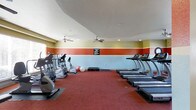 This fitness center is a dedicated single-floor space designed to meet a variety of workout needs. The room features a bold and energizing color scheme, with a tri-tone wall design incorporating shades of terracotta red, soft blue, and warm yellow, creating a motivating atmosphere for exercise. Red rubberized flooring lines the workout area, providing both durability and cushioning underfoot. The cardio section is well-appointed, offering multiple treadmills along one side of the room and a selection of elliptical cross-trainers on the opposite side, catering to users at varying fitness levels. Toward the center of the room, exercise balls and a set of free weight step platforms provide options for strength and flexibility training. Two ceiling fans are mounted overhead, promoting consistent airflow throughout the space and ensuring comfort during vigorous workouts. Recessed lighting supplements the natural light entering through windows, keeping the room bright and well-lit at all times. A posted fitness center rules board is visible on the wall, indicating a managed and maintained facility. The layout is open and spacious, allowing for ease of movement between equipment and accommodating multiple users simultaneously.