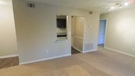 This single-floor residential space offers a practical and comfortable layout designed for everyday living. The main living area is finished with plush wall-to-wall carpeting in a warm neutral tone, creating an inviting atmosphere throughout. A distinctive pass-through window connects the living area to the kitchen, adding an open, communicative feel between the two spaces. The kitchen features dark cabinetry with granite-style countertops, offering a sleek contrast to the lighter tones of the surrounding walls. A textured ceiling adds classic architectural character to the space. Doorways lead seamlessly from the living area into additional rooms, suggesting a well-thought-out floor plan that maximizes flow and connectivity. Ventilation registers are positioned throughout, ensuring consistent climate control across the unit. The hallway beyond the main living space hints at further rooms, likely including a bedroom and additional living quarters, all finished with the same cohesive neutral palette. Natural light filters into the adjoining spaces, contributing to a bright and airy feel. Overall, the space presents a clean, functional, and well-maintained environment suited for comfortable residential living.