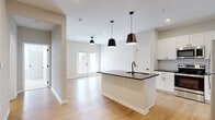 This thoughtfully designed single-floor residence offers a cohesive blend of modern aesthetics and functional living. The open-concept kitchen and dining area serves as the heart of the home, anchored by a central island with a dark granite countertop, undermount sink with a matte black faucet, and stylish pendant lighting overhead. White shaker cabinetry lines the walls, complemented by a subway tile backsplash and a full suite of stainless steel appliances, including a refrigerator, range, and microwave. Light hardwood flooring flows seamlessly throughout the main living areas, enhancing the bright, airy atmosphere. A glass-paneled door in the living area invites natural light and provides access to the outdoors. The bedroom is a comfortable retreat, finished with plush carpeting, a ceiling fan, a generous walk-in closet, and a window that brings in natural light. The bathroom features a clean, neutral palette with a dark countertop vanity, white cabinetry, matte black hardware accents, and a soaking tub with tile surround. A dedicated hallway with additional closet space adds practical storage. Throughout, the consistent use of neutral tones, clean lines, and modern fixtures creates a cohesive and inviting living environment.