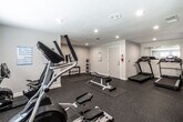 This fitness center is designed to accommodate a comprehensive range of workout routines within a thoughtfully organized, single-floor layout. The space is finished with durable rubber flooring throughout, providing cushioned support suitable for both cardio and strength training activities. Recessed lighting illuminates the area evenly, complementing natural light that enters through latticed windows along one wall.

The cardio zone features multiple treadmills equipped with individual display screens, an elliptical trainer, and a recumbent stationary bike, offering varied options for aerobic exercise. A dedicated strength training area includes a rack of dumbbells in multiple weights, a set of medicine balls, a weight bench, and additional resistance equipment. Wall-mounted workout reference charts, including dumbbell exercise and yoga pose guides, provide helpful visual instruction throughout the space.

Full-length mirrors along one wall visually expand the space while allowing users to monitor their form during exercise. A wall-mounted flat-screen television adds an entertainment option for cardio sessions. A water cooler station is conveniently positioned within the main area. Restroom facilities are accessible directly from the fitness floor. The neutral gray walls and clean white trim contribute to a functional, organized atmosphere suited for focused exercise.