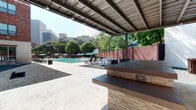 This expansive outdoor amenity space offers a thoughtfully designed environment for recreation and relaxation. At its center is a resort-style swimming pool surrounded by a spacious deck furnished with lounge chairs and teal market umbrellas, creating a vibrant, vacation-inspired atmosphere. A brick perimeter wall provides privacy while mature trees offer natural shade throughout the area.

A covered outdoor kitchen serves as a key gathering hub, featuring a stainless steel built-in grill station constructed atop stacked stone bases with dark countertops. The metal pergola overhead provides ample shade while track lighting allows for evening use. A substantial bar-height counter nearby offers additional prep and entertaining space.

Adjacent to the pool area, a dedicated game zone showcases an oversized life-size chess set with bold black-and-white checker-patterned flooring, set against a gravel landscape bed. Cornhole boards are also visible, adding to the recreational options available.

The surrounding multi-story brick building with balconies and large windows frames the courtyard, contributing an urban yet inviting character. The combination of shaded amenities, open sky, and lush tree canopy creates a dynamic and well-balanced outdoor living experience.