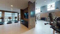 This fitness center is a thoughtfully designed, single-floor facility that combines industrial-inspired aesthetics with a full suite of exercise equipment. Exposed brick walls run throughout, paired with smooth neutral flooring, creating a warm yet energetic atmosphere. The space is divided into several functional zones, each catering to different workout styles.

The main strength training area boasts impressively high ceilings with large clerestory windows that flood the floor with natural light. Pendant light fixtures and ceiling fans complement the soaring volume of this section, which houses a comprehensive selection of free weights, dumbbell racks, weight benches, cable machines, and multi-functional strength equipment. Full-length mirrors expand the sense of space and support proper form during training.

A dedicated cardio zone lines one wall with a row of treadmills, ellipticals, and stationary bikes, positioned alongside oversized windows that offer views of the surrounding greenery outdoors. A flat-screen television mounted on a brick column provides entertainment for cardio sessions.

A separate group fitness or activity room is accessible through black-framed glass double doors, offering colorful dumbbells and a barre rail, suitable for stretching, yoga, or group classes. A water fountain is conveniently located near the entrance. The overall design balances raw industrial character with modern fitness functionality.