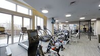This fitness center offers a comprehensive workout environment designed to accommodate a wide range of exercise routines. The space is bathed in natural light, courtesy of large windows that line the walls and create an open, energizing atmosphere throughout. The ceiling features recessed circular light fixtures that provide consistent, even illumination across the entire floor.

The cardio section is prominently featured and well-stocked, housing a variety of treadmills, elliptical trainers, stair climbers, and stationary cycling bikes, each equipped with individual digital display screens for interactive training. The equipment is arranged in an organized, accessible layout that encourages ease of movement through the space.

Toward the rear, a dedicated strength training zone offers a selection of resistance machines and free weight equipment, catering to both beginner and advanced fitness needs. Full-length mirrors line the walls in this area, enhancing the sense of depth while allowing users to monitor their form during workouts.

The flooring consists of durable, wood-toned paneling suited for high-traffic fitness use. Wall-mounted televisions provide entertainment and motivation throughout. The overall design strikes a balance between functionality and comfort, making this a well-appointed facility for a thorough and effective fitness experience.