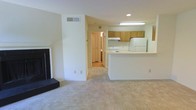 This single-floor residence offers a practical and comfortable layout centered around an open-concept living and kitchen area. The living room is anchored by a classic fireplace with a dark surround and raised hearth, providing a focal point for the space. Light-toned carpeting extends throughout the main living area, contributing to a clean and neutral aesthetic.

The kitchen is positioned adjacent to the living room and is defined by a white peninsula counter that serves as a natural divider between the two spaces. Wood-toned upper cabinetry lines the kitchen walls, complemented by a white range and refrigerator. A fluorescent overhead light fixture ensures the kitchen is well-lit and functional.

A hallway visible from the main living area leads to additional rooms, with what appears to be wood or laminate flooring in the connecting spaces, offering a contrast to the carpeted main area. Recessed lighting and flush-mounted fixtures provide consistent illumination throughout.

The overall interior palette is light and neutral, with white walls and ceilings that enhance the sense of openness. The layout flows efficiently, making the space approachable and easy to navigate. Ventilation is supported by ceiling vents, and ample wall outlets are distributed throughout for everyday convenience.