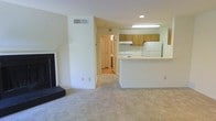 This single-floor residence offers a practical and comfortable layout centered around an open-concept living and kitchen area. The living room is anchored by a classic fireplace with a dark surround and raised hearth, providing a focal point for the space. Light-toned carpeting extends throughout the main living area, contributing to a clean and neutral aesthetic.

The kitchen is positioned adjacent to the living room and is defined by a white peninsula counter that serves as a natural divider between the two spaces. Wood-toned upper cabinetry lines the kitchen walls, complemented by a white range and refrigerator. A fluorescent overhead light fixture ensures the kitchen is well-lit and functional.

A hallway visible from the main living area leads to additional rooms, with what appears to be wood or laminate flooring in the connecting spaces, offering a contrast to the carpeted main area. Recessed lighting and flush-mounted fixtures provide consistent illumination throughout.

The overall interior palette is light and neutral, with white walls and ceilings that enhance the sense of openness. The layout flows efficiently, making the space approachable and easy to navigate. Ventilation is supported by ceiling vents, and ample wall outlets are distributed throughout for everyday convenience.