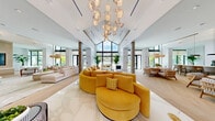Located at 15705 SW 127th Avenue in Miami, FL, this expansive single-floor clubhouse delivers an impressive blend of tropical elegance and contemporary design. The grand entrance greets visitors with large-format polished marble-look flooring, a dramatic sculptural pendant light, and an open corridor framed by floor-to-ceiling glass doors that flood the space with natural light and offer views of lush palm-lined outdoor surroundings.

The living room is a standout centerpiece, anchored by soaring vaulted ceilings clad in warm wood paneling and illuminated by a striking sculptural chandelier. A bold, curved mustard-yellow sectional sofa commands attention against a backdrop of large-scale, vibrant tropical murals that infuse the space with color and artistic energy. Additional seating areas transition seamlessly through the open-plan layout, featuring herringbone light wood flooring, woven rattan accents, and oversized windows that frame views of the pool area beyond.

A café-style lounge and dining area offers a bright, airy gathering space with pendant lighting, a long service counter, and casual seating arrangements ideal for socializing. A secondary lounge area with vaulted wood ceilings, natural fiber pendant lights, and floor-to-ceiling glazing extends the indoor-outdoor connection. Throughout, the design maintains a cohesive coastal-modern aesthetic, enriched by curated artwork, lush indoor greenery, and an abundance of natural light.