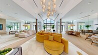 Located at 15705 SW 127th Avenue in Miami, FL, this expansive single-floor clubhouse delivers an impressive blend of tropical elegance and contemporary design. The grand entrance greets visitors with large-format polished marble-look flooring, a dramatic sculptural pendant light, and an open corridor framed by floor-to-ceiling glass doors that flood the space with natural light and offer views of lush palm-lined outdoor surroundings.

The living room is a standout centerpiece, anchored by soaring vaulted ceilings clad in warm wood paneling and illuminated by a striking sculptural chandelier. A bold, curved mustard-yellow sectional sofa commands attention against a backdrop of large-scale, vibrant tropical murals that infuse the space with color and artistic energy. Additional seating areas transition seamlessly through the open-plan layout, featuring herringbone light wood flooring, woven rattan accents, and oversized windows that frame views of the pool area beyond.

A café-style lounge and dining area offers a bright, airy gathering space with pendant lighting, a long service counter, and casual seating arrangements ideal for socializing. A secondary lounge area with vaulted wood ceilings, natural fiber pendant lights, and floor-to-ceiling glazing extends the indoor-outdoor connection. Throughout, the design maintains a cohesive coastal-modern aesthetic, enriched by curated artwork, lush indoor greenery, and an abundance of natural light.