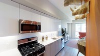 This single-floor space showcases a contemporary open-concept design that combines style and functionality. The kitchen serves as a central highlight, featuring floor-to-ceiling high-gloss white cabinetry that maximizes vertical storage while lending a clean, minimalist aesthetic. Stainless steel appliances, including a gas range and built-in microwave, offer a professional-grade cooking experience. The white countertops provide ample workspace and contrast elegantly with the warm-toned lower cabinetry. A stainless steel undermount sink is positioned beneath under-cabinet lighting, which illuminates the work surfaces effectively. White subway tile backsplash adds a classic yet modern touch to the space. Light hardwood flooring runs throughout, connecting the kitchen seamlessly to the adjacent living area. Large windows at the far end of the space allow generous amounts of natural light to filter through, brightening the interior and offering views of the outdoors. A patterned area rug and lush green plants add warmth and character to the living zone. A distinctive sculptural decorative element is visible overhead, providing a unique artistic focal point that adds personality to the otherwise clean-lined interior.