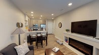 This well-appointed single-floor residence offers a thoughtfully designed open-concept layout that seamlessly connects the living, dining, and kitchen areas. The living room serves as a central gathering space, anchored by a sleek electric fireplace with built-in shelving and a large flat-screen TV mounted above. Recessed lighting illuminates the warm wood-look flooring throughout, while windows fitted with blinds allow for adjustable natural light. A comfortable seating arrangement and a wood coffee table complete the inviting atmosphere.

The kitchen is a standout feature, showcasing modern two-tone cabinetry in a warm gray finish, a classic subway tile backsplash, and a suite of stainless steel appliances including a range, over-the-range microwave, and a French door refrigerator. A peninsula with a deep single-basin sink and light countertops provides additional prep space and casual dining overflow. A dedicated dining area accommodates a full table and chair set, positioned conveniently between the kitchen and living room.

The bedroom offers a serene retreat with a ceiling fan, a window for natural light, and direct access to a generous walk-in closet outfitted with wire shelving and hanging rods. The full bathroom features tile flooring, a tub-shower combination with white tile surround, and matching gray cabinetry. In-unit laundry is also included.