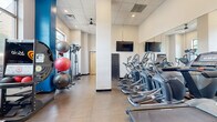 This fitness center offers a comprehensive workout environment designed to accommodate a wide range of exercise routines. The space is defined by its clean, modern aesthetic, featuring a neutral tile floor, a drop ceiling with recessed lighting, and striking blue accent columns that add a bold visual contrast to the white walls.

The cardio zone is well-stocked with a row of elliptical trainers and treadmills, including Matrix-brand machines equipped with individual display screens. Full-length wall mirrors along one side visually expand the space and allow users to monitor their form during exercise. Wall-mounted flat-screen televisions are positioned throughout, providing entertainment during workouts.

The strength and functional training area includes a multi-tier dumbbell rack with a wide range of weights, an adjustable workout bench, a resistance training machine, and a rack of stability balls in varying sizes. Additional specialty equipment, such as a solo training unit, rounds out the functional fitness offerings.

Large windows along the perimeter allow natural light to fill the space and offer views of the surrounding area. A ceiling fan provides additional ventilation, contributing to a comfortable workout environment overall.