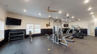 This single-level fitness center in Tontitown, Arkansas, offers a comprehensive workout environment designed for both cardio and strength training. The space features a two-tone interior design, with crisp white upper walls contrasting against dark shiplap-style wainscoting that runs along the lower half of each wall, creating a polished, modern aesthetic. Durable rubber flooring covers the entire space, providing cushioning and traction suitable for high-intensity use.

The room is well-lit with recessed ceiling lights supplemented by large ceiling fans, ensuring comfortable airflow throughout workouts. Natural light enters through windows fitted with white blinds, adding brightness to the space.

Equipment includes a multi-station cable machine by Hoist, multiple True-brand treadmills, elliptical cross-trainers, a rowing machine, and a stationary spin bike. A dedicated free weights area features a full dumbbell rack alongside an adjustable weight bench, with a wall-mounted flat-screen TV positioned nearby for entertainment. Additional stability balls are stored along the wall, rounding out the functional training options.

The layout is open and spacious, allowing users to move freely between stations without congestion. The overall design balances functionality with a clean, inviting atmosphere well-suited for regular fitness use.