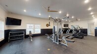 This single-level fitness center in Tontitown, Arkansas, offers a comprehensive workout environment designed for both cardio and strength training. The space features a two-tone interior design, with crisp white upper walls contrasting against dark shiplap-style wainscoting that runs along the lower half of each wall, creating a polished, modern aesthetic. Durable rubber flooring covers the entire space, providing cushioning and traction suitable for high-intensity use.

The room is well-lit with recessed ceiling lights supplemented by large ceiling fans, ensuring comfortable airflow throughout workouts. Natural light enters through windows fitted with white blinds, adding brightness to the space.

Equipment includes a multi-station cable machine by Hoist, multiple True-brand treadmills, elliptical cross-trainers, a rowing machine, and a stationary spin bike. A dedicated free weights area features a full dumbbell rack alongside an adjustable weight bench, with a wall-mounted flat-screen TV positioned nearby for entertainment. Additional stability balls are stored along the wall, rounding out the functional training options.

The layout is open and spacious, allowing users to move freely between stations without congestion. The overall design balances functionality with a clean, inviting atmosphere well-suited for regular fitness use.