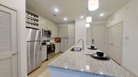This single-floor residence is designed with a clean, contemporary aesthetic that emphasizes functionality and modern finishes throughout. The kitchen serves as a central highlight, featuring white shaker-style cabinetry, granite countertops, and a decorative arabesque tile backsplash. A spacious island with an undermount stainless steel sink and pendant lighting above provides both a prep area and casual dining space. Stainless steel appliances, including a refrigerator, range, and microwave, complete the culinary setup. A built-in wine rack adds a practical design touch.

The open layout connects the kitchen seamlessly to the living area, which benefits from natural light via floor-to-ceiling glass doors leading to a balcony. A ceiling fan, light wood-style flooring, and neutral tones throughout contribute to an airy, cohesive atmosphere.

The bedrooms are carpeted for comfort, each featuring ceiling fans, natural light from large windows, and ample space for furnishings. The bathroom is finished with granite countertops, a dual-sink vanity with modern cabinetry, and light wood flooring. A walk-in closet with built-in hanging rods connects directly to the bathroom, offering generous storage.
