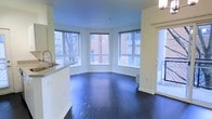 This single-floor space showcases a clean, modern design with an open-concept layout that maximizes both functionality and visual appeal. The standout feature of the living area is a striking curved bay window configuration, with multiple floor-to-ceiling windows arranged in a sweeping arc. This architectural detail floods the space with abundant natural light and offers views of the surrounding neighborhood, including mature trees and adjacent brick buildings. Rich, dark hardwood floors run throughout, creating a bold contrast against the crisp white walls and trim, lending the space a refined yet contemporary character. The open-plan kitchen is positioned adjacent to the living area, featuring a light-toned countertop and a modern sink with a sleek pull-down faucet. The kitchen peninsula provides a natural visual boundary between the cooking and living zones while maintaining the open, airy feel of the space. A glass-paneled door is visible along one wall, offering access to an exterior area and bringing in additional natural light. White baseboards and smooth ceilings complete the polished interior aesthetic. Overall, the space presents a well-balanced combination of architectural interest, natural light, and modern finishes that create a welcoming and functional environment.